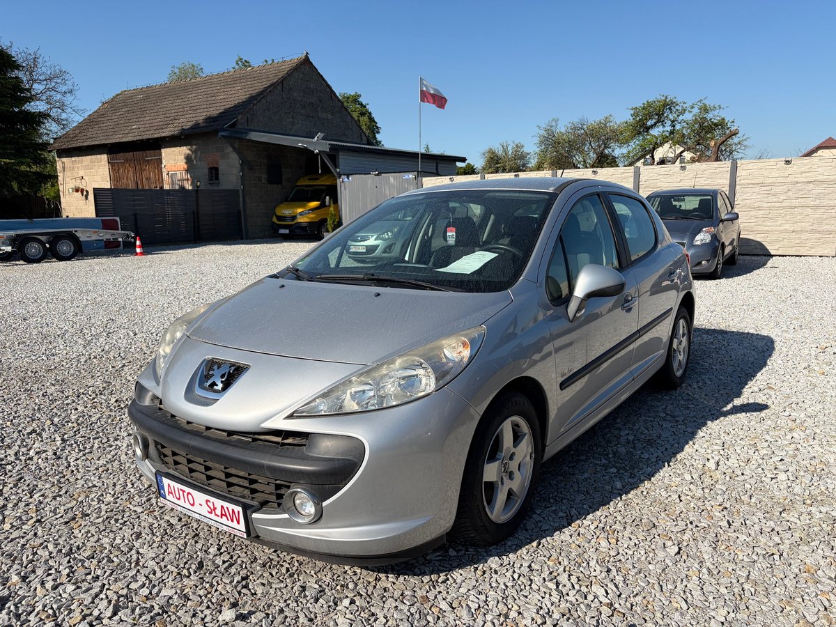 Silver Peugeot 207 sedan parked at a car dealership with a brick building and flag in the background