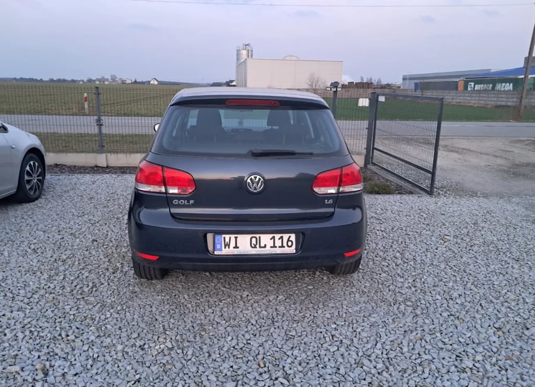 Rear view of a dark gray Volkswagen Golf parked on a gravel lot with industrial buildings in the background