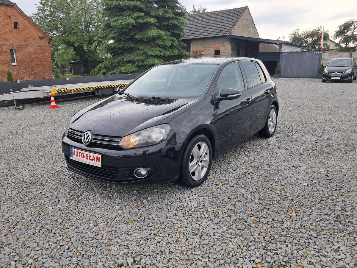 Black Volkswagen Golf parked on gravel lot with brick buildings and barrels in background