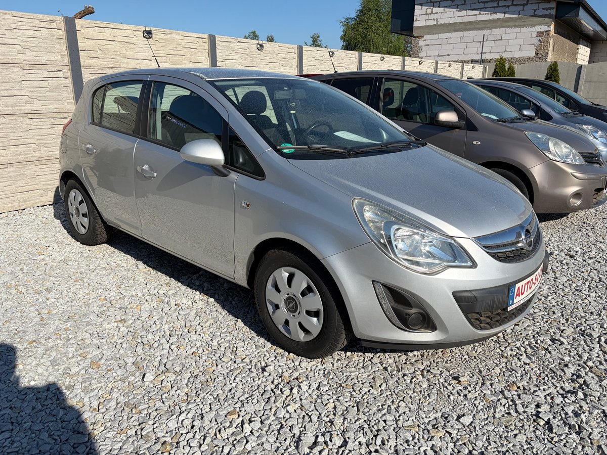 Silver Opel Corsa compact car parked in gravel lot with beige fence and another car in background
