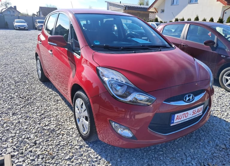 Red Hyundai i10 compact car parked in a gravel lot with residential buildings in background