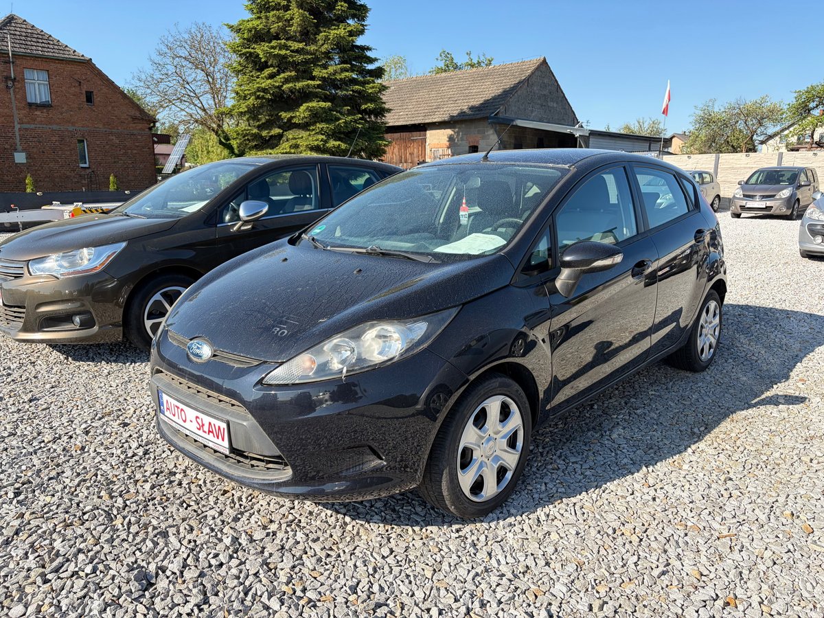 Black Ford Fiesta parked on gravel in a car dealership lot with other vehicles and buildings in background