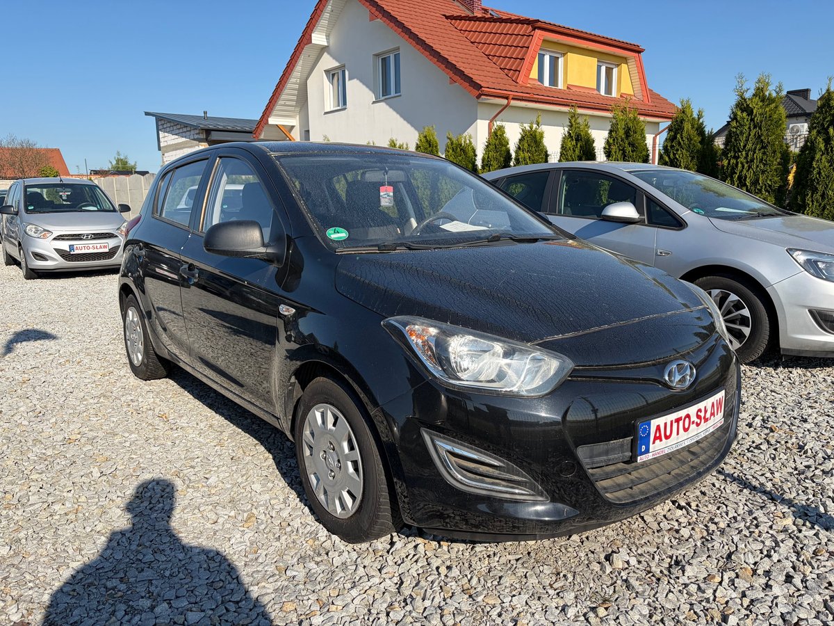 Black Hyundai i20 hatchback car parked in gravel driveway with red-roofed house and other vehicles in background