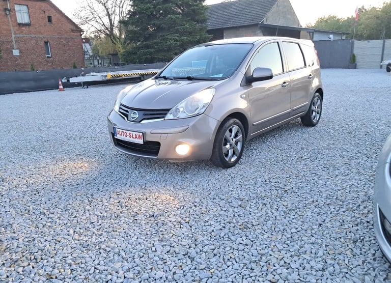 Silver Nissan Note compact car parked on gravel driveway with brick building and trees in background