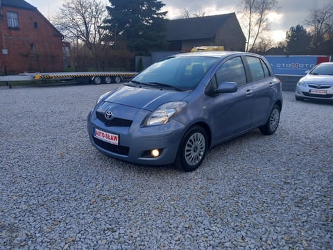 Blue Toyota Yaris parked on gravel lot with brick building and cars in background